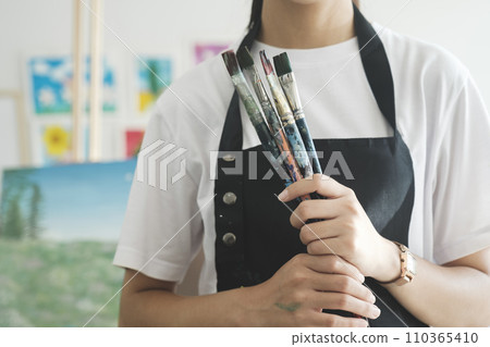 A close-up of the artist hands wearing an apron smeared with paint. Clutching many brushes and paintbrushes. A close-up of the artist hands wearing an apron smeared with paint. Clutching many brushes and paintbrushes. 110365410