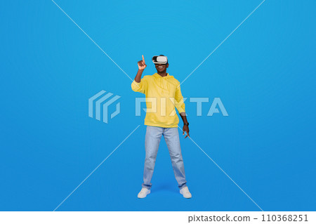 A Black man in a yellow hoodie points upwards while wearing virtual reality goggles 110368251