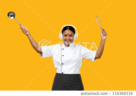 Smiling black female chef dancing with kitchen utensils in hands Smiling black female chef dancing with kitchen utensils in hands 110368316