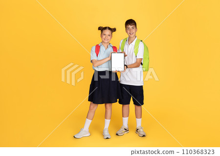 A smiling boy and girl in school uniforms, with backpacks, stand side by side holding a tablet 110368323