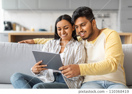 Happy Young Indian Couple Using Digital Tablet While Relaxing On Couch 110368419