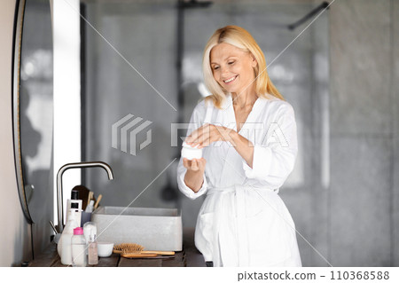 Smiling senior woman in white bathrobe opening jar of face cream 110368588