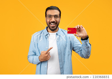 A cheerful man in a denim shirt holding a red credit card and pointing at it with his other hand A cheerful man in a denim shirt holding a red credit card and pointing at it with his other hand 110368831