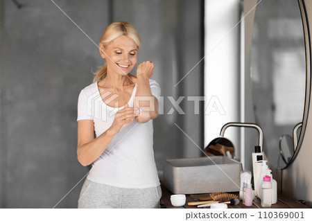 Smiling mature woman in casual clothes applying body lotion to her elbow Smiling mature woman in casual clothes applying body lotion to her elbow 110369001
