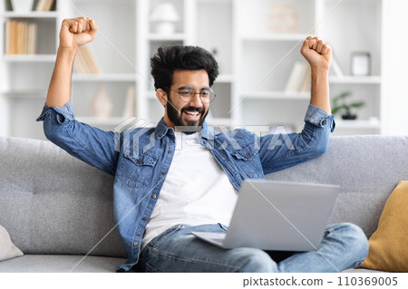 Joyful indian man raising fists in victory while looking at laptop screen 110369005