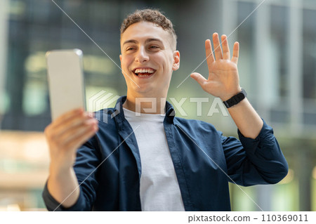 Joyful young man with short curly hair video calling on his smartphone 110369011