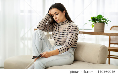 A contemplative young woman in a striped top and jeans lounges on a sofa, phone in hand A contemplative young woman in a striped top and jeans lounges on a sofa, phone in hand 110369016