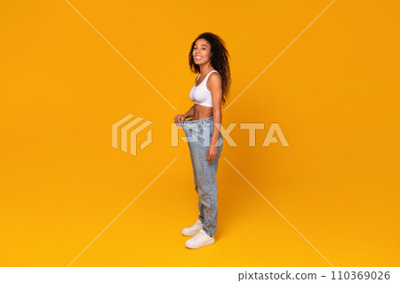 Happy black woman demonstrating weight loss wearing oversized pants, studio 110369026