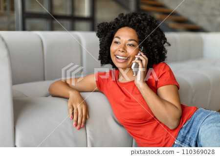 Cheerful young black woman chatting on her smartphone while relaxing at home Cheerful young black woman chatting on her smartphone while relaxing at home 110369028