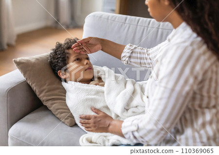 Caring black mother checking her son's forehead while he lying on couch Caring black mother checking her son's forehead while he lying on couch 110369065