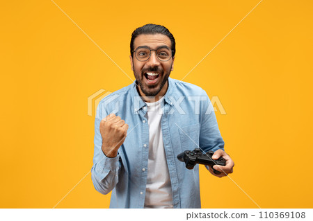 An overjoyed man with a beard and glasses holding a game controller clenches his fist An overjoyed man with a beard and glasses holding a game controller clenches his fist 110369108