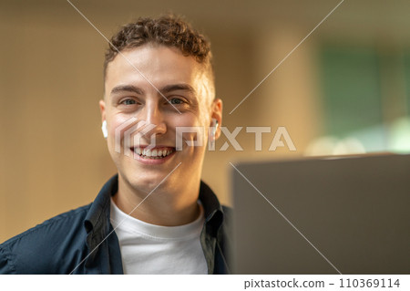 A young man with a beaming smile, using earbuds, and looking at a laptop screen 110369114