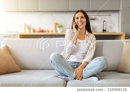 Happy young woman chatting on cellphone, sitting cross-legged on sofa at home Happy young woman chatting on cellphone, sitting cross-legged on sofa at home 110369116