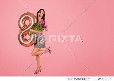 Joyful woman in a shimmering silver dress exuberantly holds a rose gold number eight balloon 110369297