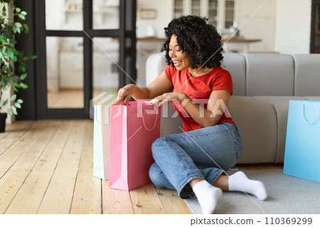 Happy young black woman sitting on floor, looking into colorful shopping bags 110369299