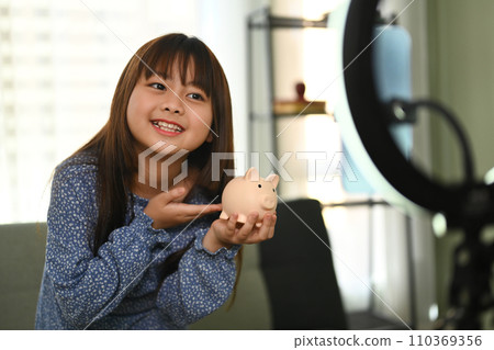 Pretty asian girl holding piggybank in front of camera while recording video on smartphone. Saving money for future concept 110369356