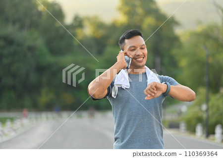 Smiling sportsman having pleasant phone conversation and checking time on watch while exercise in the park Smiling sportsman having pleasant phone conversation and checking time on watch while exercise in the park 110369364