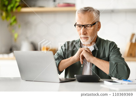 Thoughtful elderly man wearing glasses using laptop and calculator at home Thoughtful elderly man wearing glasses using laptop and calculator at home 110369369