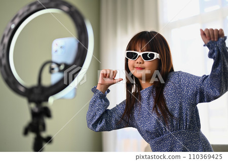 Smiling Asian girl dancing on smartphone camera at home while filming for social media Smiling Asian girl dancing on smartphone camera at home while filming for social media 110369425