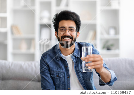 Cheerful young Indian man in denim jacket holding glass of water Cheerful young Indian man in denim jacket holding glass of water 110369427