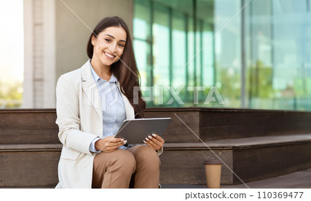 Cheerful young professional woman sitting on a bench with a digital tablet, smartwatch on her wrist Cheerful young professional woman sitting on a bench with a digital tablet, smartwatch on her wrist 110369477