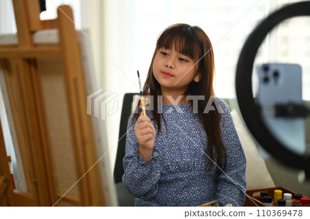 Cute Asian schoolgirl painting picture and recording video on smartphone at home. 110369478