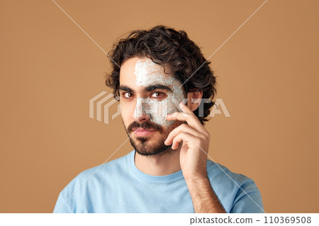 Calm brunette with curly hair, bearded man with dermatology facial care mask and looking at camera against beige studio background. 110369508