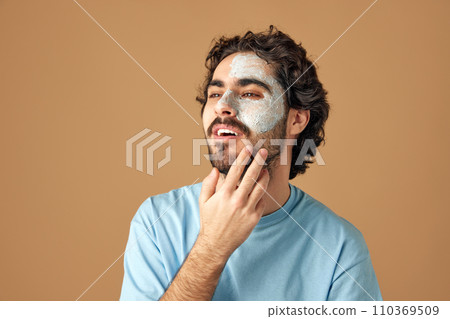 Unshaved brunette man with curly hair applies facial care mask against beige studio background. Dermatology health, luxury spa procedures 110369509