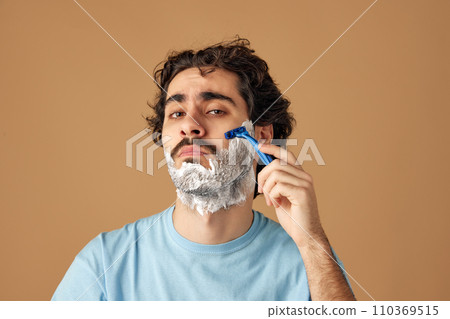 Portrait of young brunette man with curly hair applies shaving beard against sandy studio background. Hygiene. Portrait of young brunette man with curly hair applies shaving beard against sandy studio background. Hygiene. 110369515