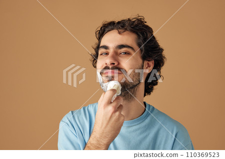 Portrait of young brunette man with curly hair applies shaving foam with retro shaving brush against sandy studio background. Portrait of young brunette man with curly hair applies shaving foam with retro shaving brush against sandy studio background. 110369523