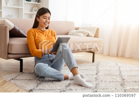 Relaxed young korean woman sitting on carpet, using digital tablet 110369597