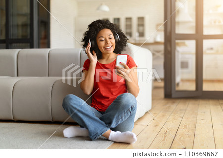 Cheerful young black woman sitting on floor, listening music on her smartphone 110369667