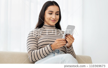Glad young arab woman typing by smartphone at sofa, watch video, read message Glad young arab woman typing by smartphone at sofa, watch video, read message 110369673
