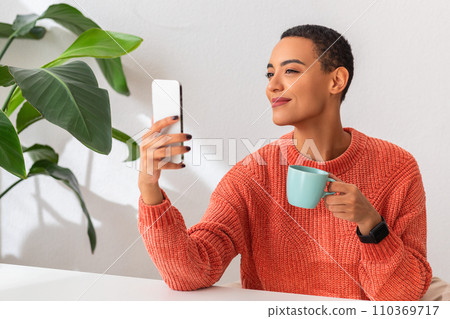 Smiling woman in an orange sweater enjoys a moment of leisure, holding a cup Smiling woman in an orange sweater enjoys a moment of leisure, holding a cup 110369717