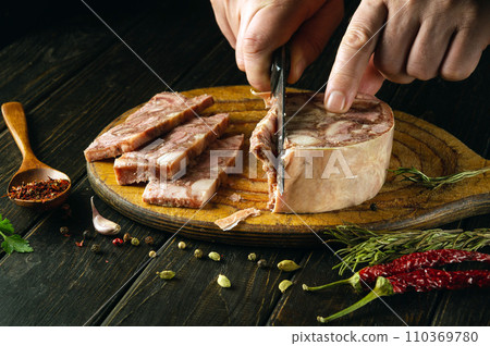 Close-up of a man with a knife in his hand cutting headcheese on the kitchen table to prepare the Belgian national dish. Peasant food Close-up of a man with a knife in his hand cutting headcheese on the kitchen table to prepare the Belgian national dish. Peasant food 110369780