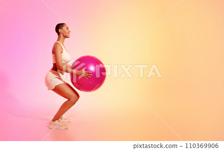 A fit woman with a shaved head performs a squat exercise with a pink fitness ball 110369906