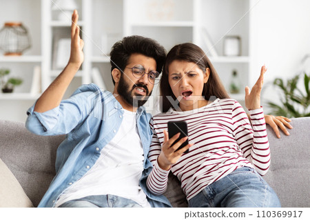 Furious indian couple looking at smartphone screen, home interior Furious indian couple looking at smartphone screen, home interior 110369917