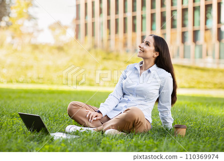 Smiling serene professional arab millennial lady sits with laptop in grass outdoor Smiling serene professional arab millennial lady sits with laptop in grass outdoor 110369974