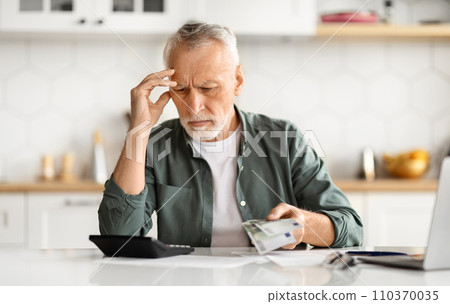 Anxious senior man managing finances, holding cash and using calculator at home Anxious senior man managing finances, holding cash and using calculator at home 110370035