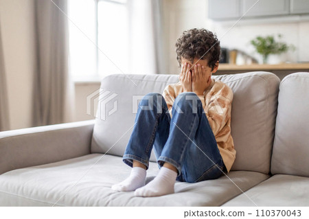 Distressed little black boy sitting on sofa, covering his face with hands 110370043