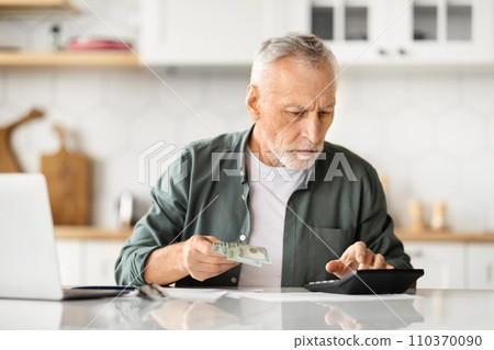 Focused elderly man holding money cash and using calculator at home 110370090