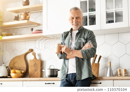 Cheerful elderly gentleman standing with mug of coffee in hands in kitchen 110370104