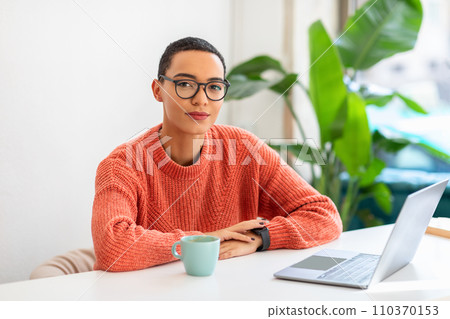 Confident professional woman with glasses sits at a white table with a mug and laptop Confident professional woman with glasses sits at a white table with a mug and laptop 110370153