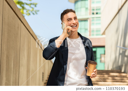 A young man in a casual outfit with a jacket, walking outdoors, talking on a mobile phone 110370193
