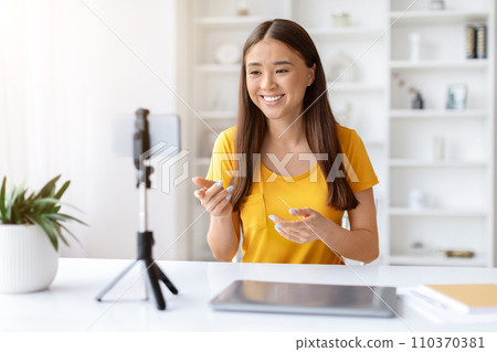 Enthusiastic asian woman vlogging at home, speaking to smartphone on tripod 110370381