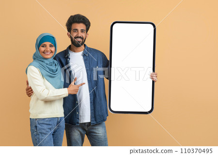 Mobile Offer. Young Muslim Couple Pointing At Blank Smartphone In Their Hands Mobile Offer. Young Muslim Couple Pointing At Blank Smartphone In Their Hands 110370495