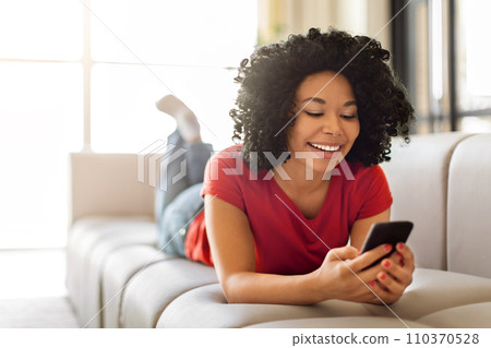 Happy young black woman messaging on smartphone while relaxing at home Happy young black woman messaging on smartphone while relaxing at home 110370528