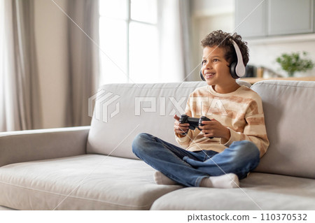 Engrossed black little boy in headphones using game controller at home Engrossed black little boy in headphones using game controller at home 110370532