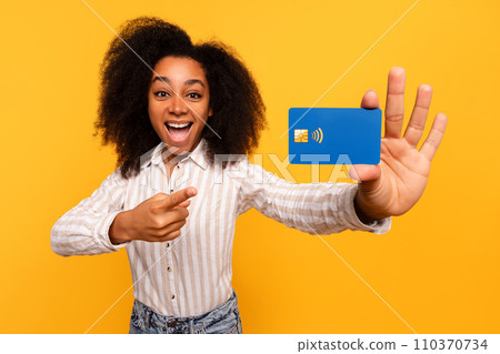 Joyful black woman presenting blue credit card, vibrant yellow backdrop 110370734