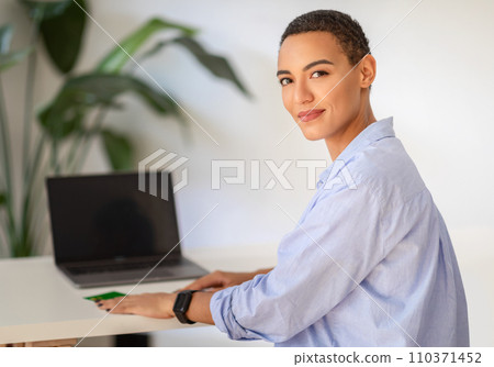 A professional young woman with short hair confidently poses by a desk with a laptop A professional young woman with short hair confidently poses by a desk with a laptop 110371452